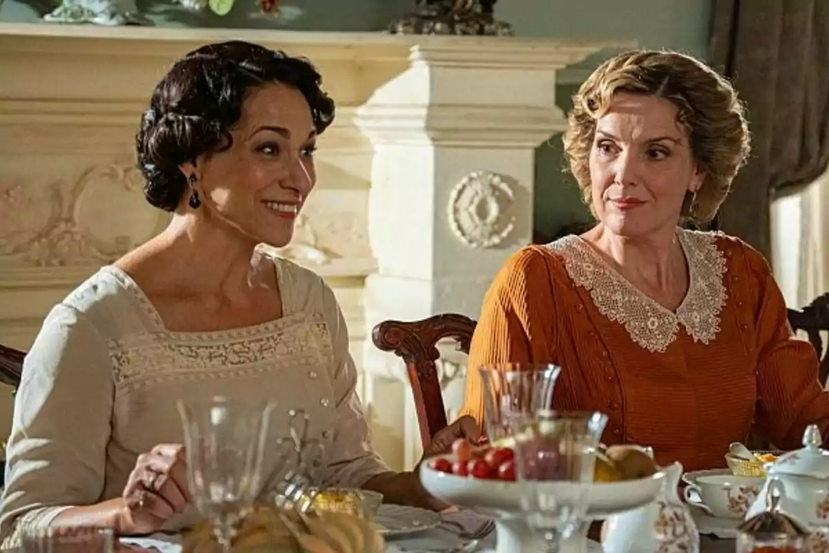Dos mujeres sentadas a la mesa en una escena de época, que son Eva Martín e Isabel Serrano, una de ellas sonríe mientras la otra la observa con atención, rodeadas de vajilla y comida. Dos mujeres sentadas a la mesa en una escena de época, que son Eva Martín e Isabel Serrano, una de ellas sonríe mientras la otra la observa con atención, rodeadas de vajilla y comida.