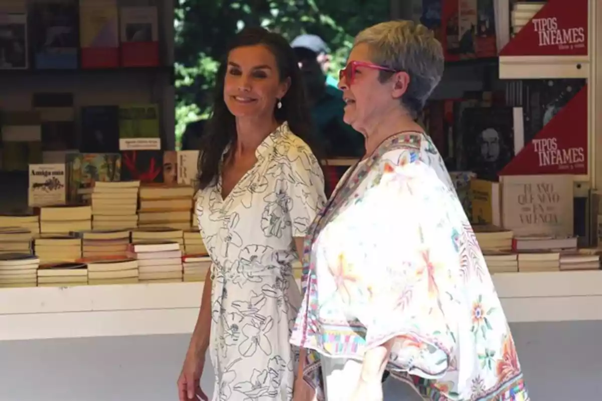 Dos mujeres sonríen mientras caminan frente a un puesto de libros en una feria al aire libre Dos mujeres sonríen mientras caminan frente a un puesto de libros en una feria al aire libre