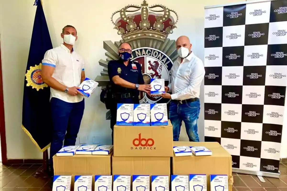 Tres personas con mascarillas posan junto a cajas de mascarillas frente a un escudo de la Policía Nacional y un banner de Spain Consulting Group.