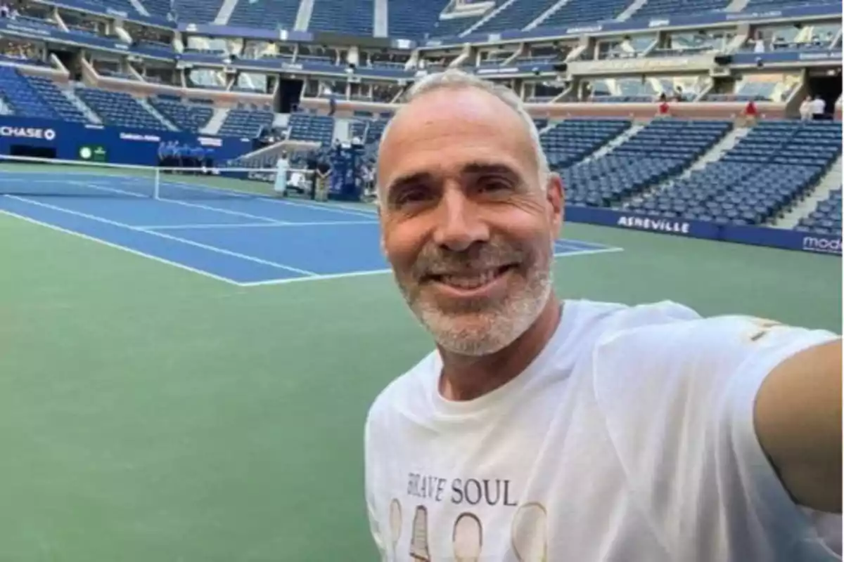 Un hombre con barba y cabello canoso sonríe mientras se toma una selfie en una cancha de tenis vacía con gradas azules al fondo.