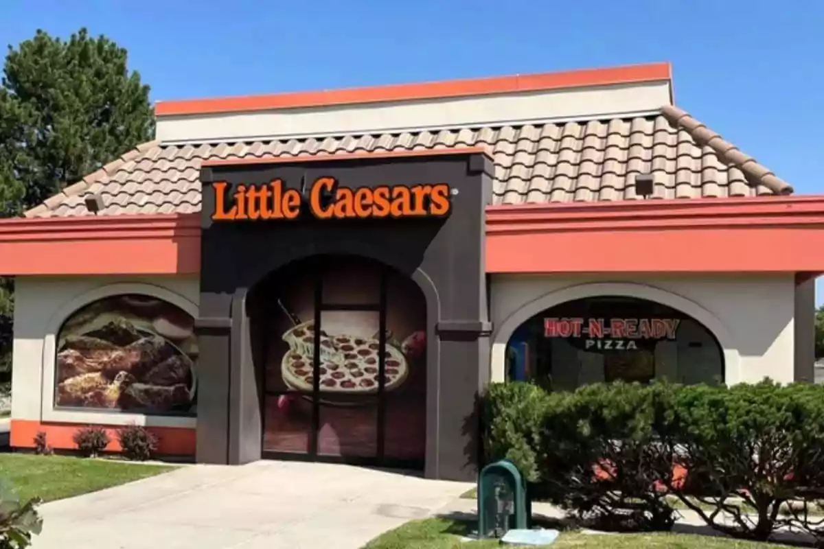 Frente de una pizzería con letrero naranja y negro, ventanas decoradas con imágenes de pizza y arbustos en el exterior. Frente de una pizzería con letrero naranja y negro, ventanas decoradas con imágenes de pizza y arbustos en el exterior.