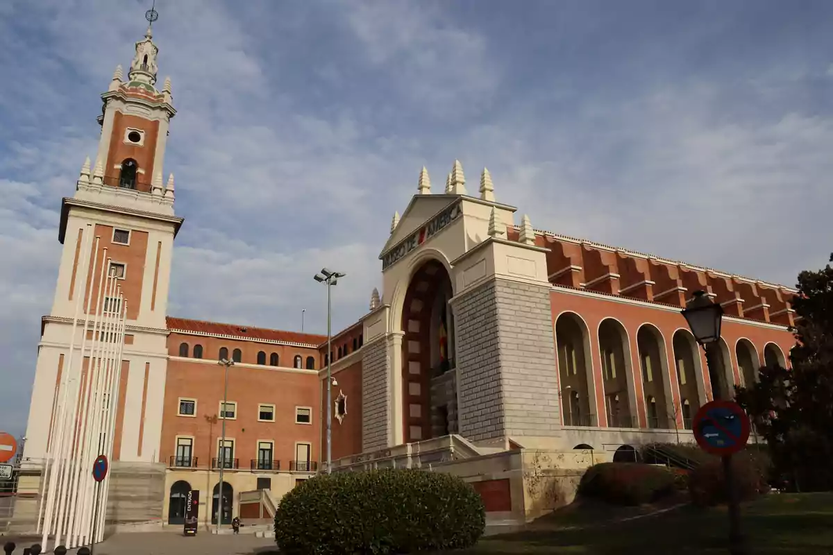 Edificio del Museo de América en Madrid con una torre alta y una fachada de ladrillo rojo bajo un cielo parcialmente nublado.