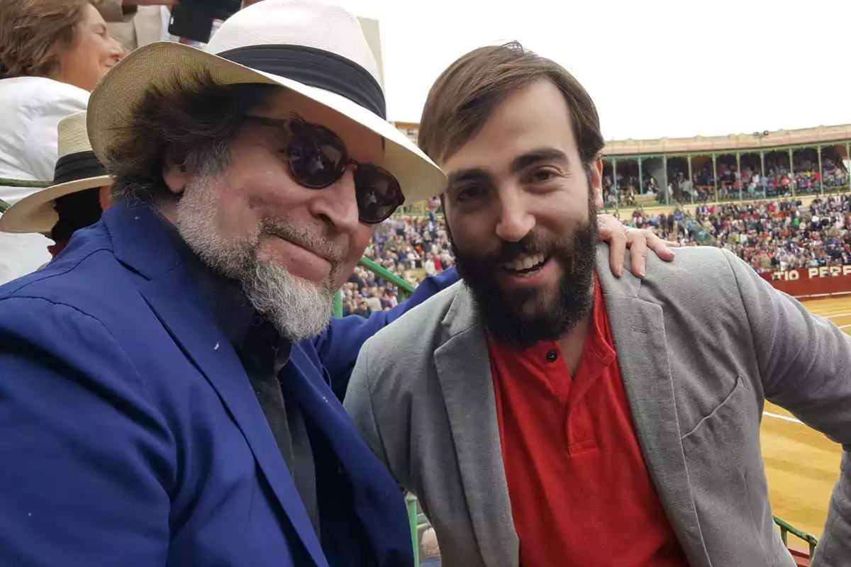 Dos personas sonríen mientras posan para una foto en un evento al aire libre, una de ellas lleva un sombrero y gafas de sol.