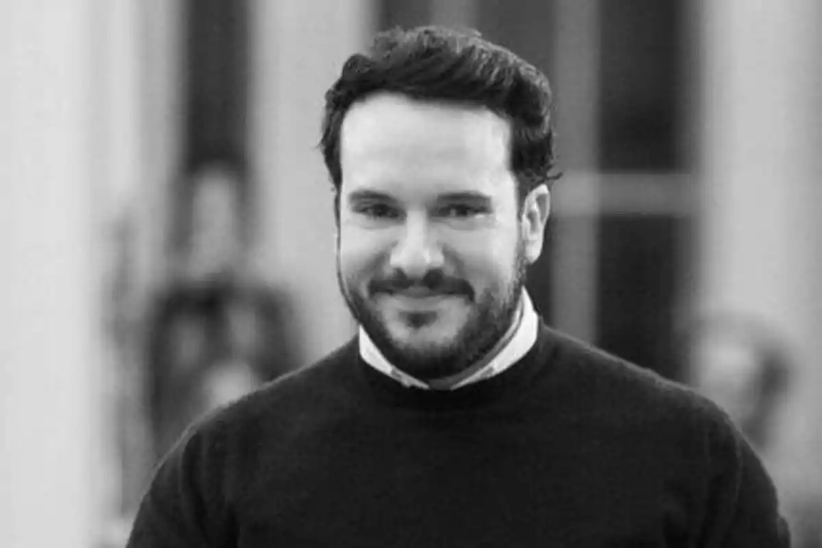 Un hombre con barba y cabello oscuro sonríe mientras mira a la cámara en una fotografía en blanco y negro.
