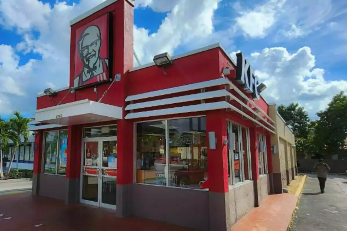 Un restaurante de comida rápida con fachada roja y un letrero grande en la parte superior -que es KFC-, rodeado de un cielo azul con nubes. Un restaurante de comida rápida con fachada roja y un letrero grande en la parte superior -que es KFC-, rodeado de un cielo azul con nubes.