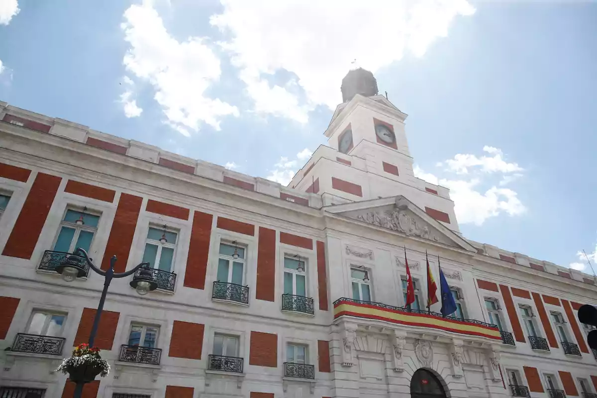 Edificio histórico con fachada de ladrillo y piedra blanca, con un reloj en la torre central y varias banderas ondeando en el balcón.