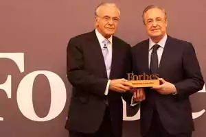 Dos hombres de traje sosteniendo un trofeo de Forbes frente a un fondo con el logo de Forbes.