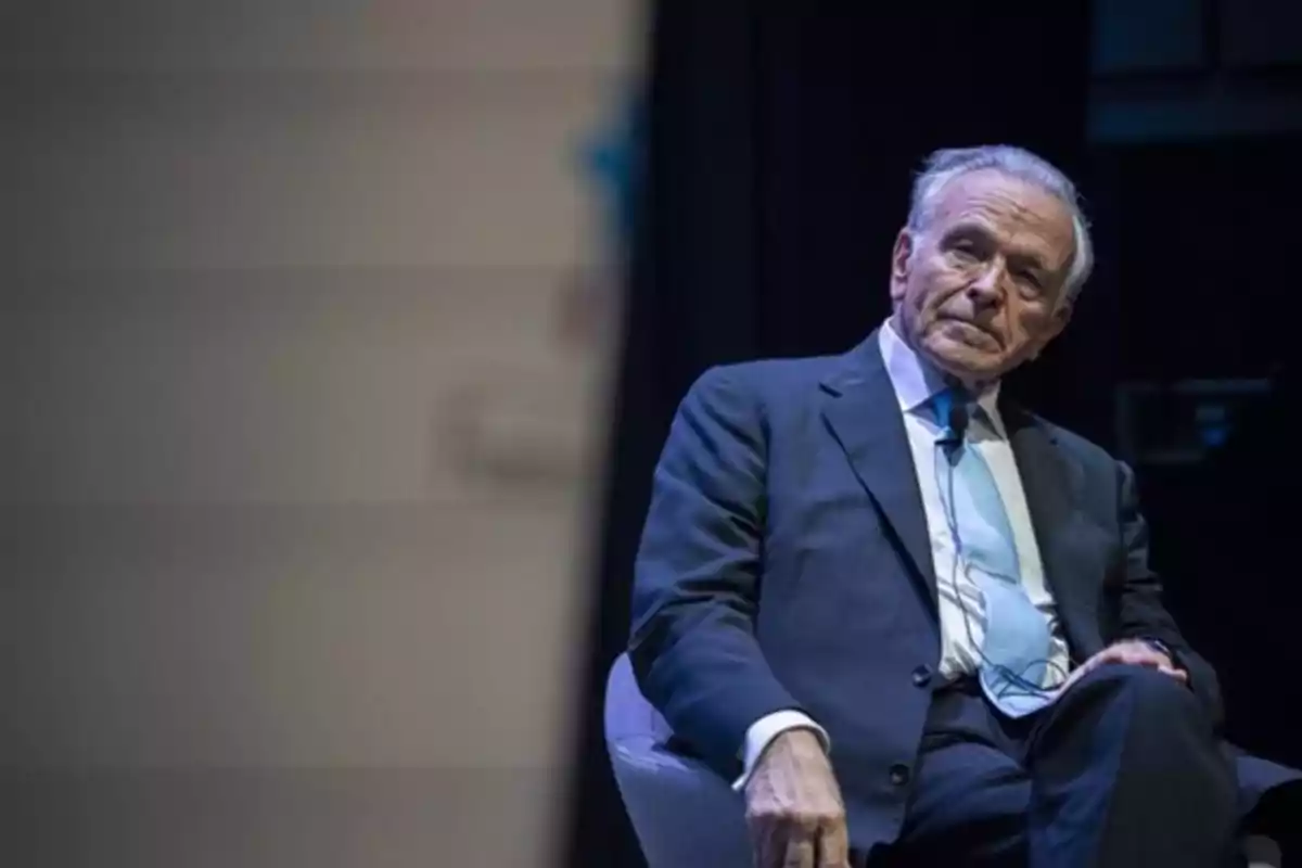 Un hombre mayor con traje oscuro y corbata azul claro está sentado en una silla durante un evento o conferencia. Un hombre mayor con traje oscuro y corbata azul claro está sentado en una silla durante un evento o conferencia.