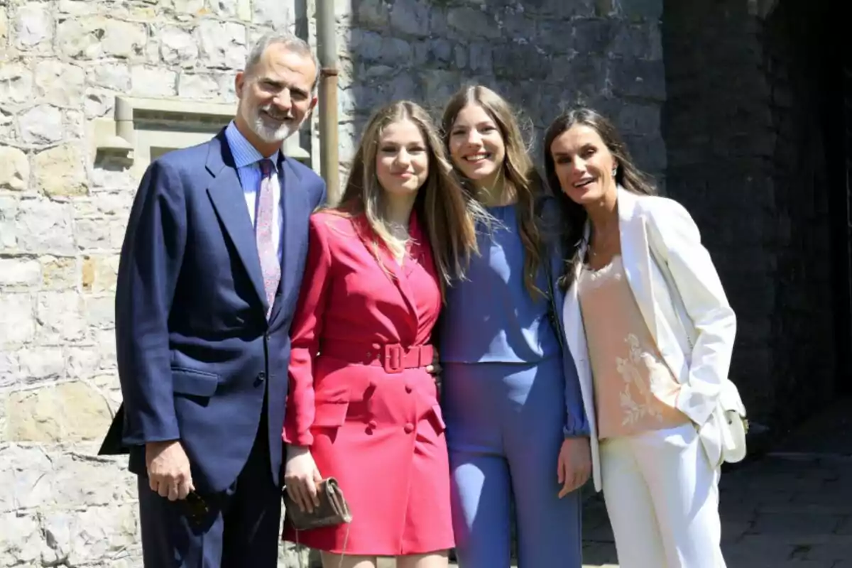 Un grupo de cuatro personas posando juntas frente a una pared de piedra, dos de ellas visten trajes formales y las otras dos llevan conjuntos de colores brillantes. Un grupo de cuatro personas posando juntas frente a una pared de piedra, dos de ellas visten trajes formales y las otras dos llevan conjuntos de colores brillantes.