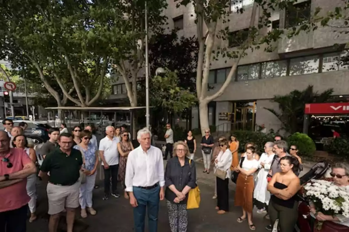 Un grupo de personas se encuentra reunido en una calle arbolada frente a un edificio.