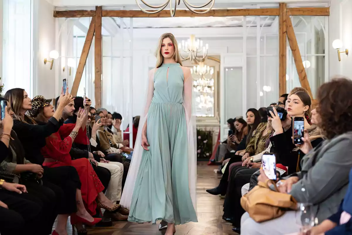 Una modelo camina por la pasarela con un vestido largo y fluido de color azul claro mientras el público observa y toma fotos en un evento de moda elegante. Una modelo camina por la pasarela con un vestido largo y fluido de color azul claro mientras el público observa y toma fotos en un evento de moda elegante.