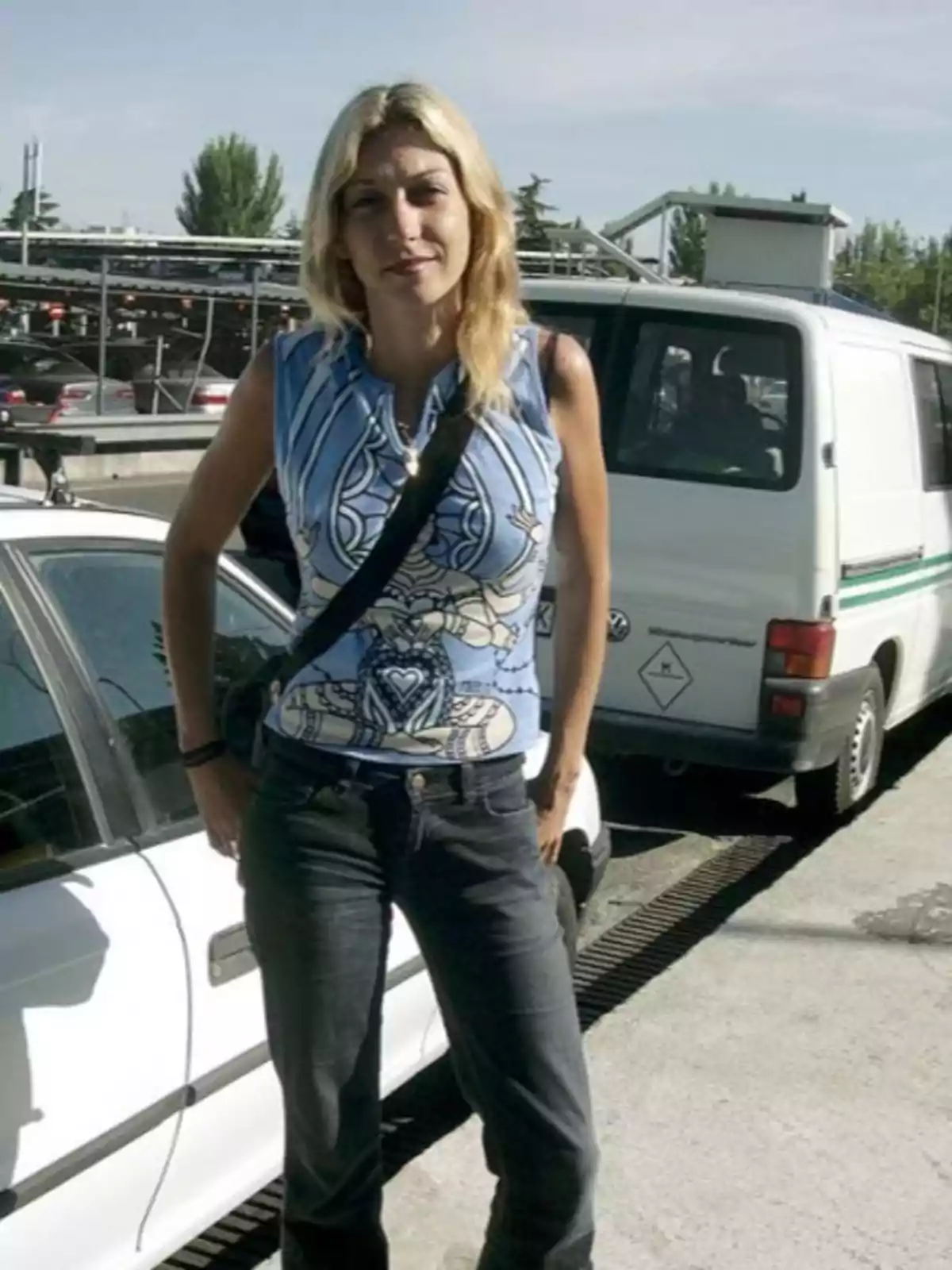 Una mujer rubia con una camiseta azul y jeans oscuros está de pie junto a un coche blanco en un aparcamiento al aire libre.