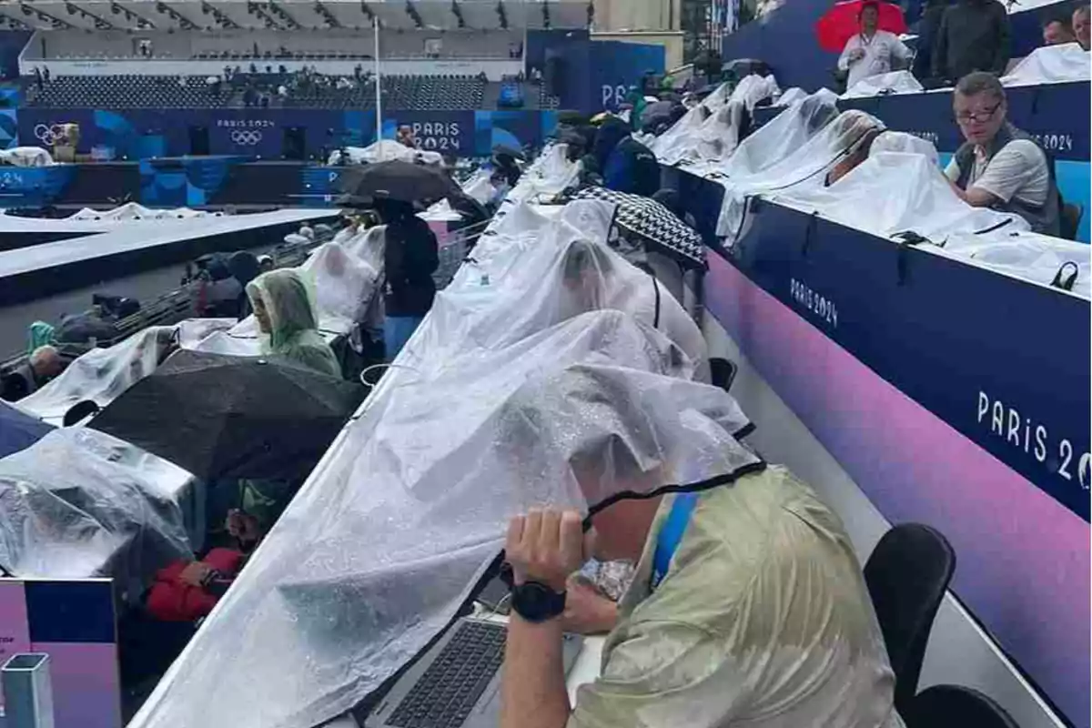 Personas bajo la lluvia en un evento deportivo en París 2024, cubiertas con plásticos y paraguas.