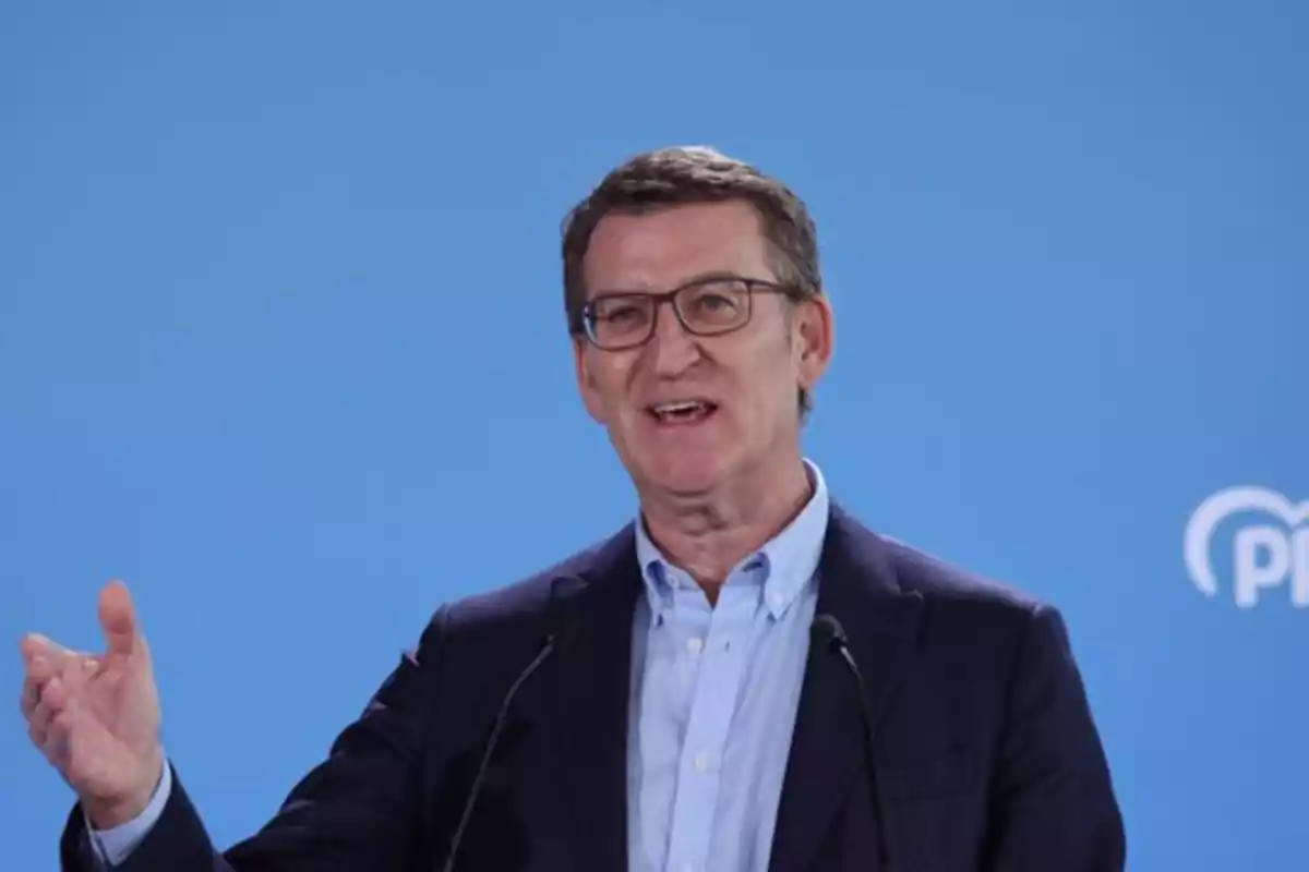 Un hombre con gafas y traje oscuro hablando frente a un micrófono con un fondo azul. Un hombre con gafas y traje oscuro hablando frente a un micrófono con un fondo azul.