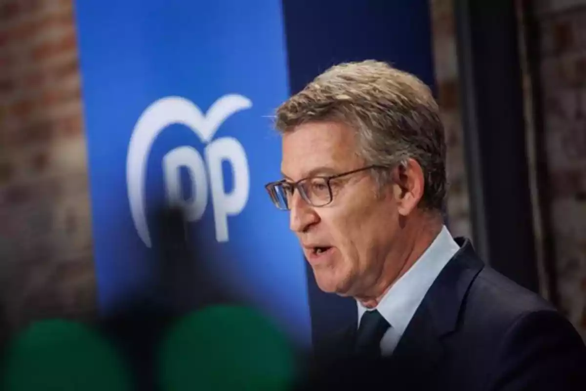 Hombre de cabello canoso y gafas hablando frente a un cartel azul con el logo del Partido Popular Hombre de cabello canoso y gafas hablando frente a un cartel azul con el logo del Partido Popular