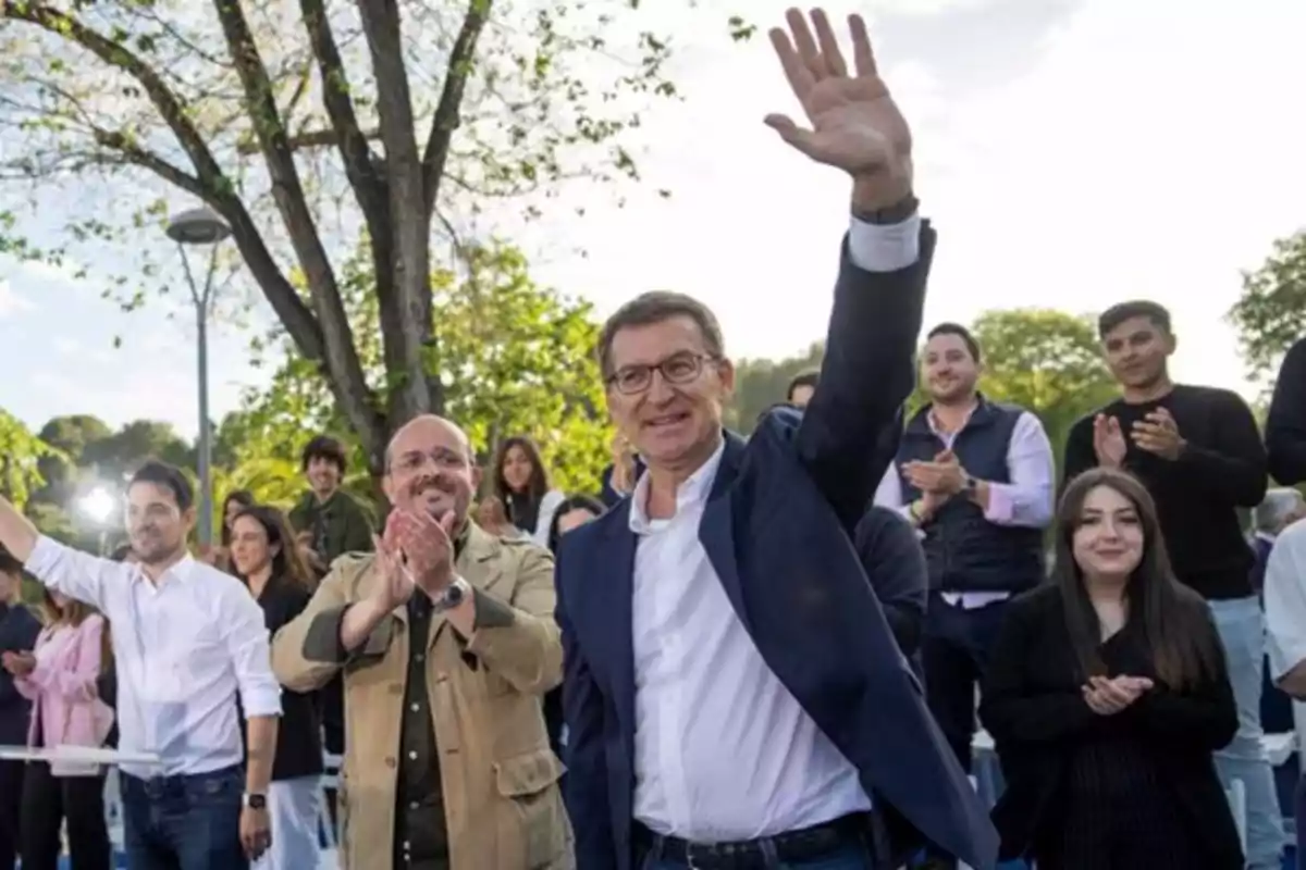 Un hombre con gafas y chaqueta azul saluda con la mano levantada mientras varias personas a su alrededor aplauden al aire libre Un hombre con gafas y chaqueta azul saluda con la mano levantada mientras varias personas a su alrededor aplauden al aire libre