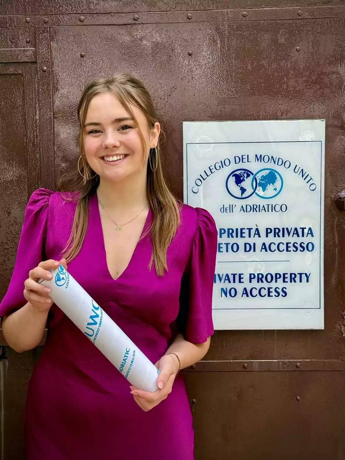 Una persona sonriente con un vestido morado sostiene un diploma frente a un cartel que indica propiedad privada y acceso restringido en el Collegio del Mondo Unito dell'Adriatico. Una persona sonriente con un vestido morado sostiene un diploma frente a un cartel que indica propiedad privada y acceso restringido en el Collegio del Mondo Unito dell'Adriatico.