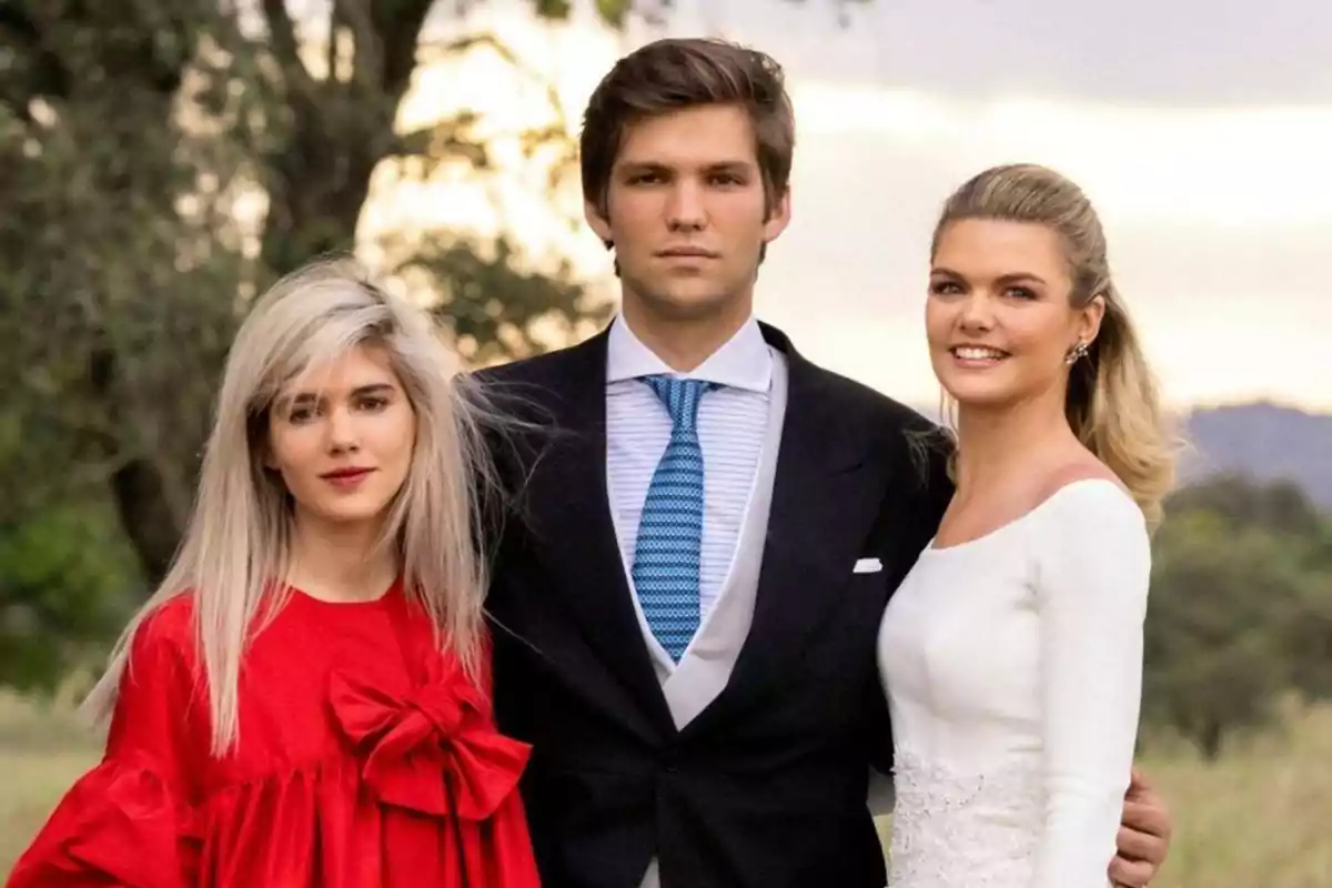 Tres personas posando juntas al aire libre, una mujer con vestido rojo, un hombre con traje y corbata azul, y otra mujer con vestido blanco.