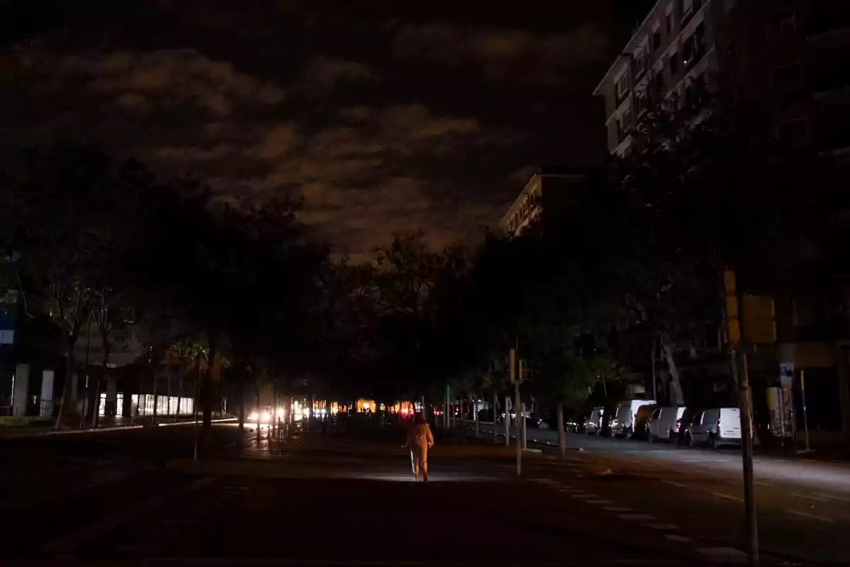 Una persona camina por una calle oscura iluminada por luces de vehículos con edificios y árboles a los lados y un cielo nublado arriba. Una persona camina por una calle oscura iluminada por luces de vehículos con edificios y árboles a los lados y un cielo nublado arriba.