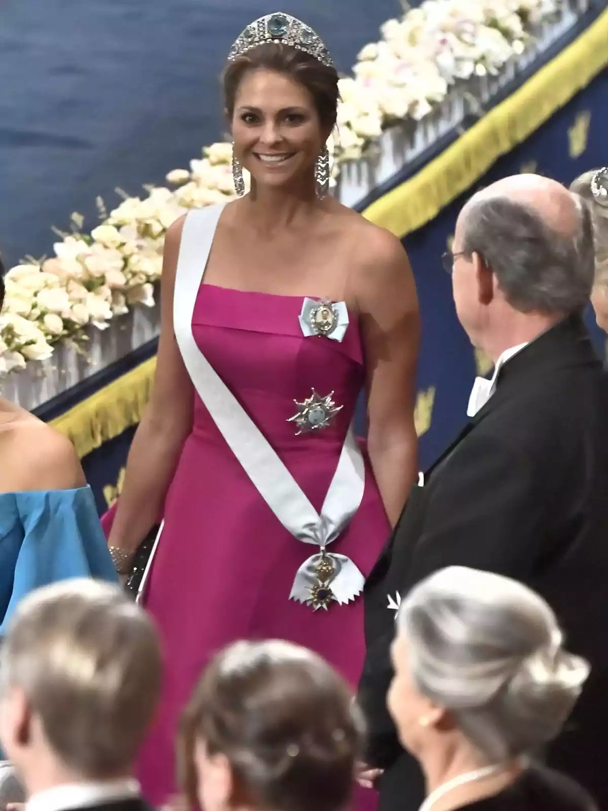 Mujer con vestido fucsia y tiara sonríe en un evento formal rodeada de otras personas elegantemente vestidas Mujer con vestido fucsia y tiara sonríe en un evento formal rodeada de otras personas elegantemente vestidas