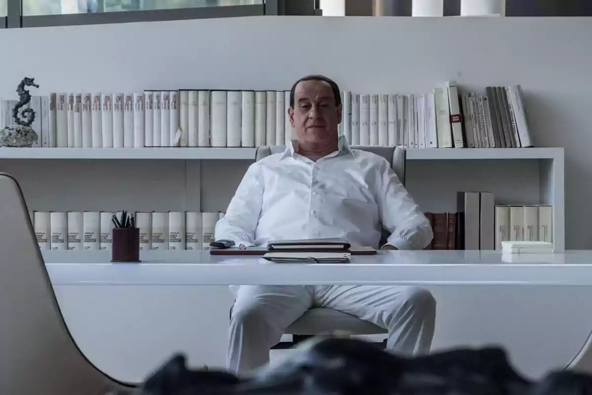 Hombre sentado en un escritorio blanco con estanterías llenas de libros detrás de él.