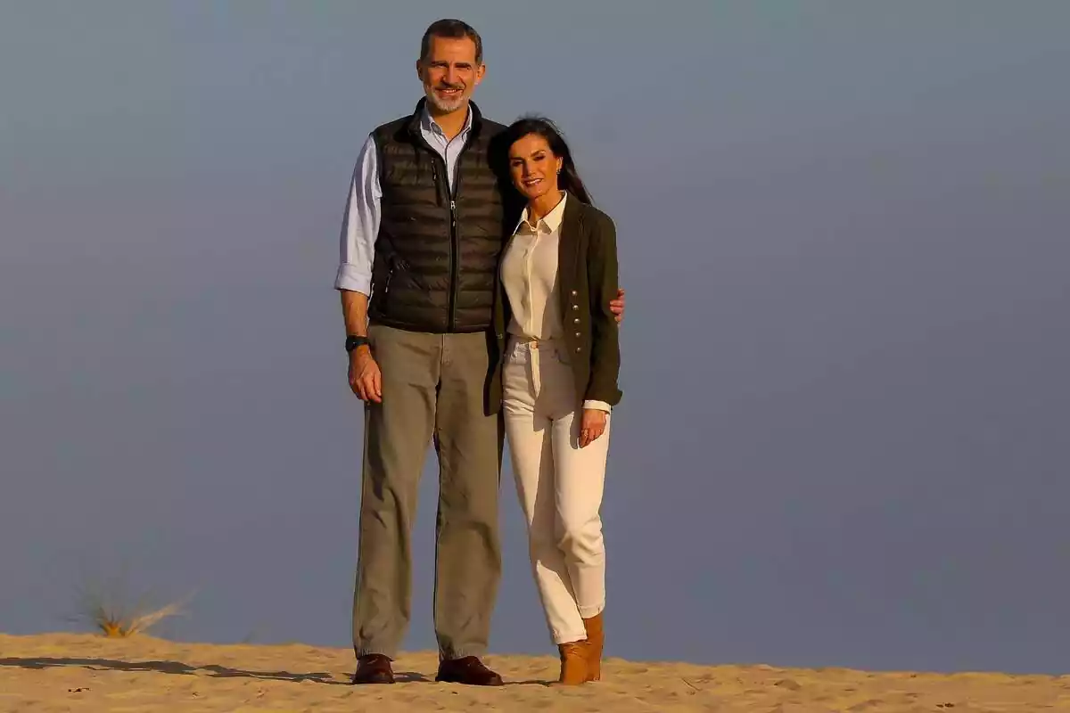 Felipe VI sonriendo con chaleco negro abrazando por al espalda a Letizia sonriendo con blazer verde y vestida de blanco en la arena Felipe VI sonriendo con chaleco negro abrazando por al espalda a Letizia sonriendo con blazer verde y vestida de blanco en la arena