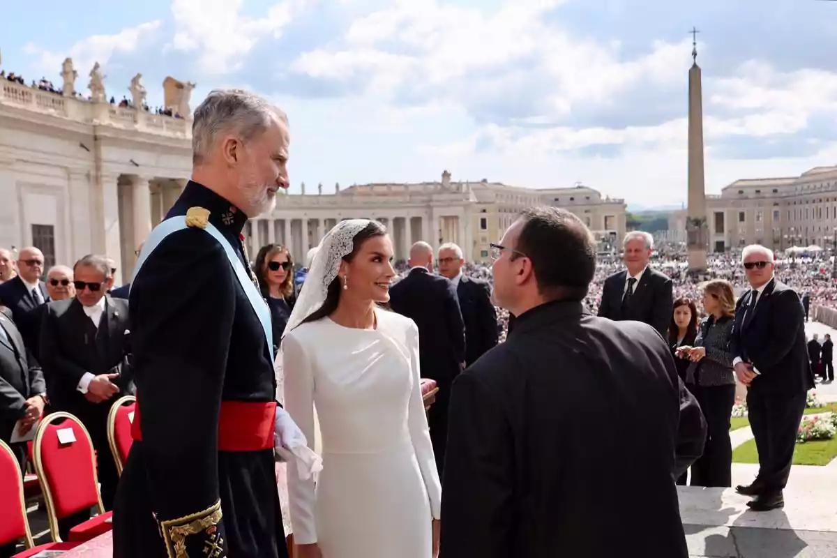 Felipe y Letizia elegantemente vestidos conversan con un sacerdote en una plaza concurrida con arquitectura clásica de fondo. Felipe y Letizia elegantemente vestidos conversan con un sacerdote en una plaza concurrida con arquitectura clásica de fondo.