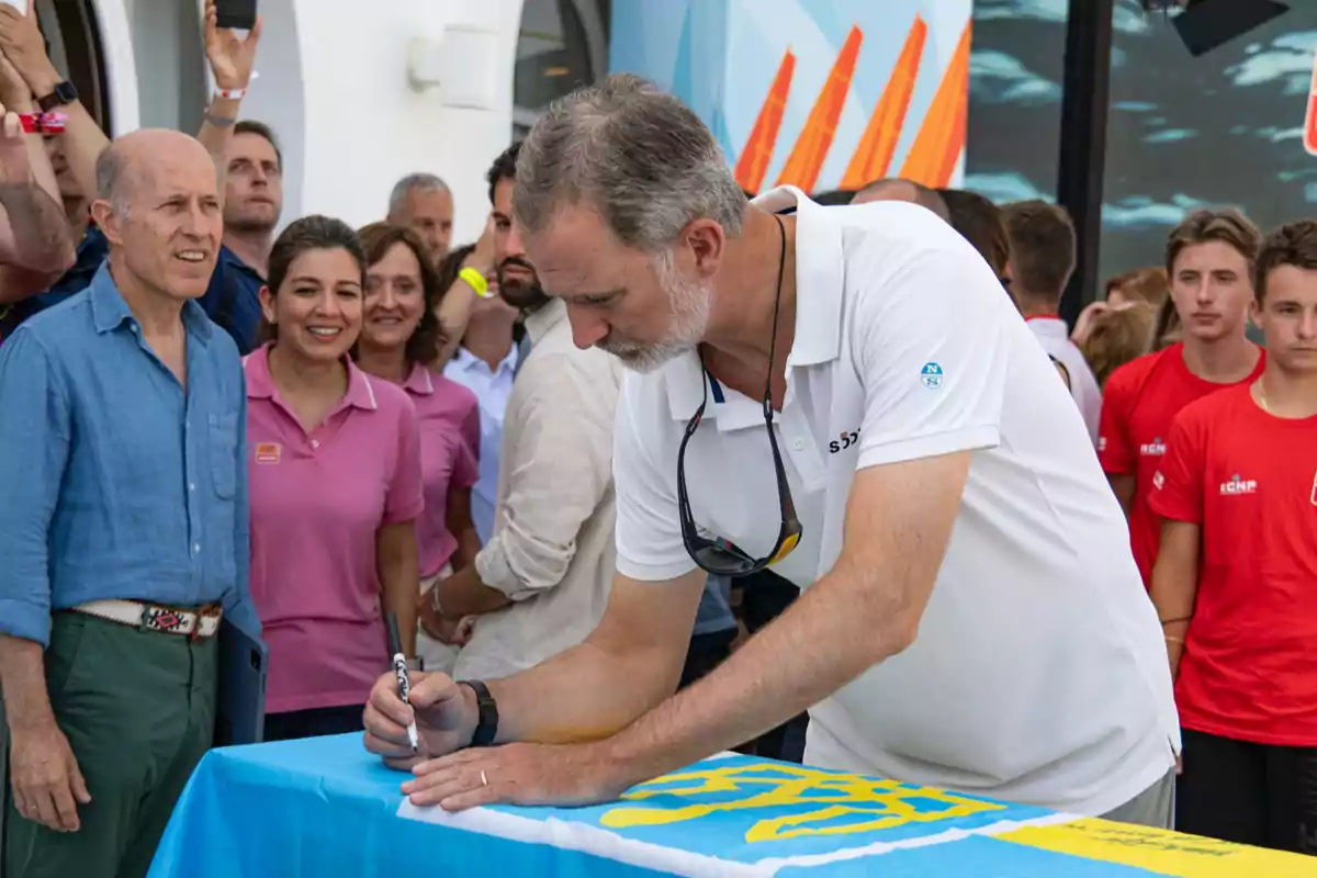 Un hombre con barba y cabello canoso, vestido con una camiseta blanca, firma un objeto mientras varias personas lo observan y algunas toman fotos.