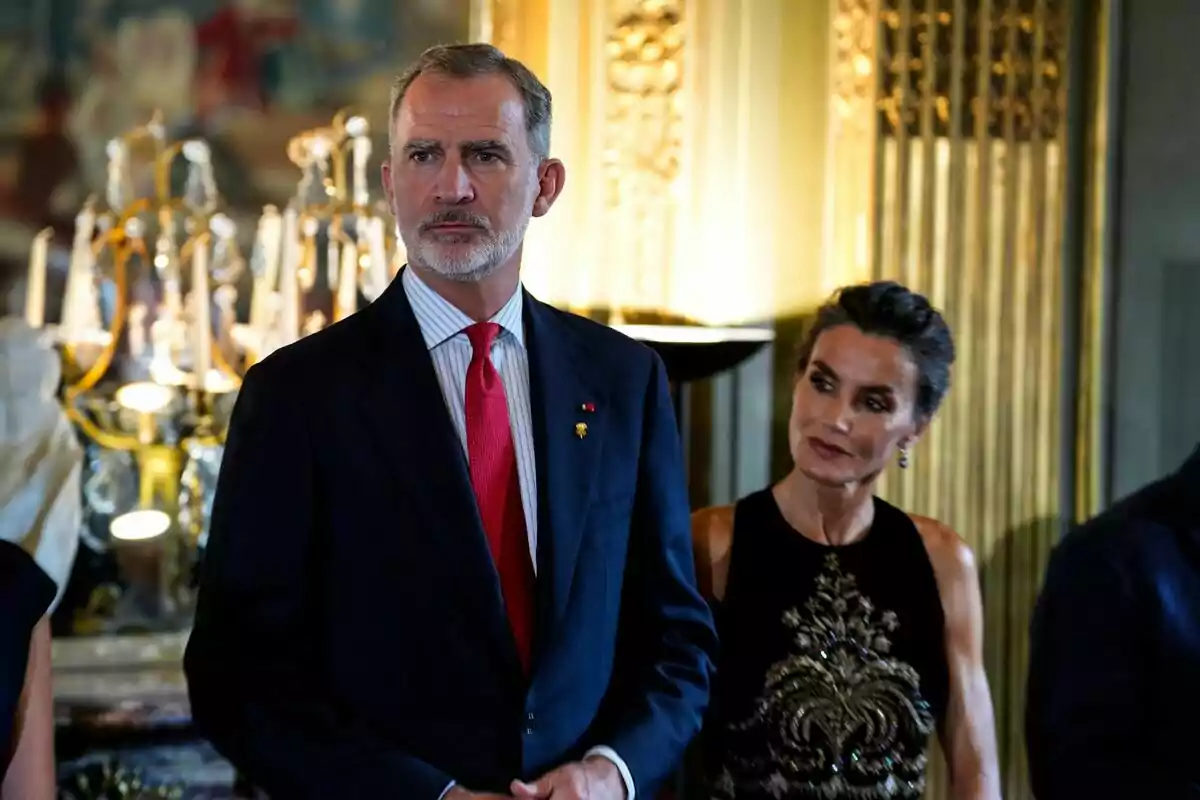 Felipe VI y Letizia elegantemente vestidos en un evento formal con un candelabro de fondo.