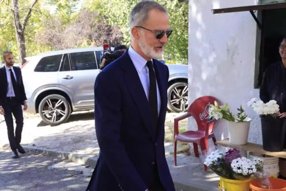 Un hombre con traje oscuro y gafas de sol camina al aire libre, mientras otra persona en traje lo sigue; al fondo hay un coche plateado y una silla roja con flores en macetas.