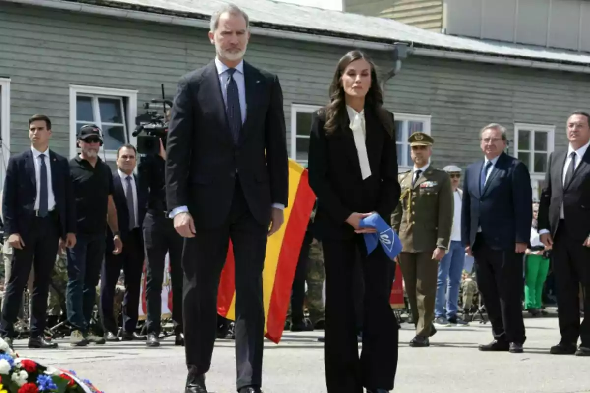 Un grupo de personas vestidas formalmente participa en una ceremonia al aire libre con una bandera española y personal militar presente. Un grupo de personas vestidas formalmente participa en una ceremonia al aire libre con una bandera española y personal militar presente.