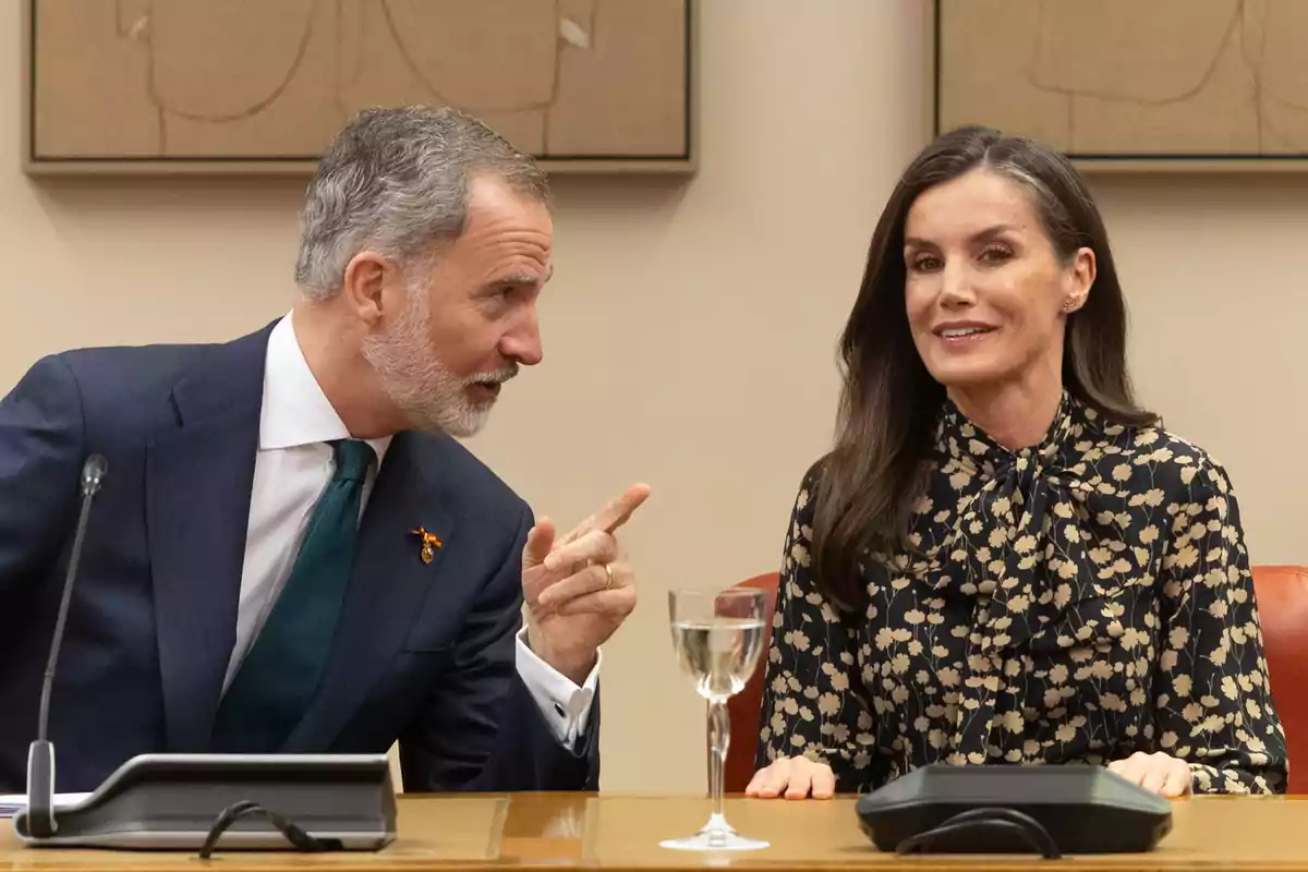 Felipe VI y la reina Letizia sentadas en una mesa conversando, una de ellas gesticula mientras la otra sonríe, con un micrófono y una copa de agua frente a ellos. Felipe VI y la reina Letizia sentadas en una mesa conversando, una de ellas gesticula mientras la otra sonríe, con un micrófono y una copa de agua frente a ellos.