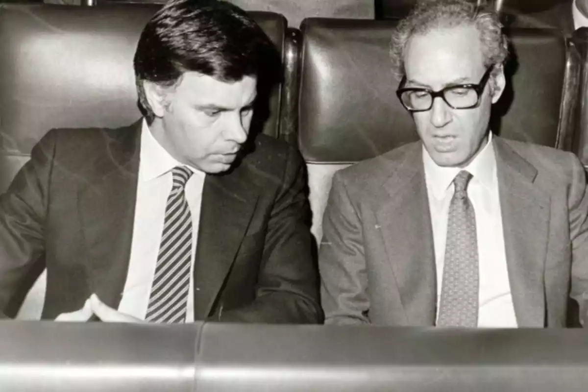 El expresidente Felipe González, sentado al lado del exministro Miguel Boyer. Ambos están hablando y mirando a un punto que hay entre ambos encima de una mesa.