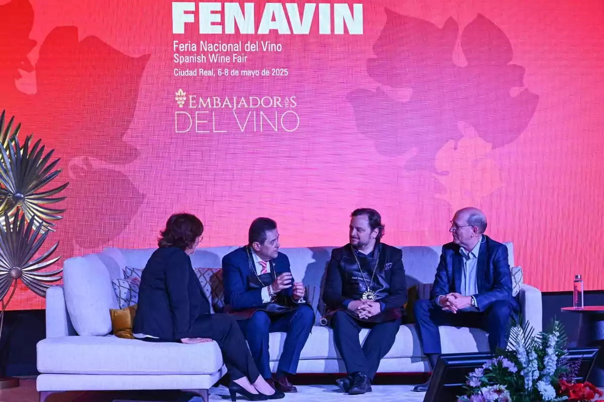 Personas sentadas en un sofá durante un evento de la Feria Nacional del Vino con un fondo rojo que muestra el texto Personas sentadas en un sofá durante un evento de la Feria Nacional del Vino con un fondo rojo que muestra el texto