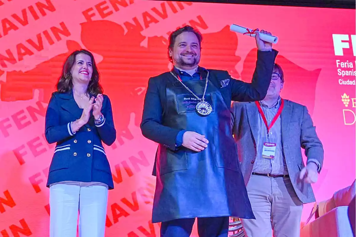 Un hombre sonriente sostiene un diploma mientras una mujer aplaude y otro hombre está de pie a su lado en un evento con un fondo rojo que dice "FENAVIN".