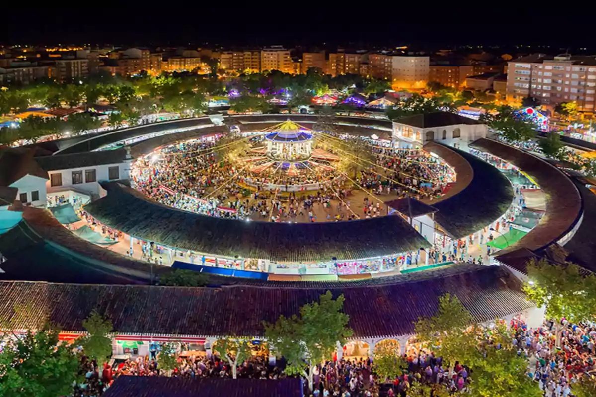 Vista aérea de una feria nocturna con una estructura circular iluminada y llena de gente, rodeada de árboles y edificios en el fondo.