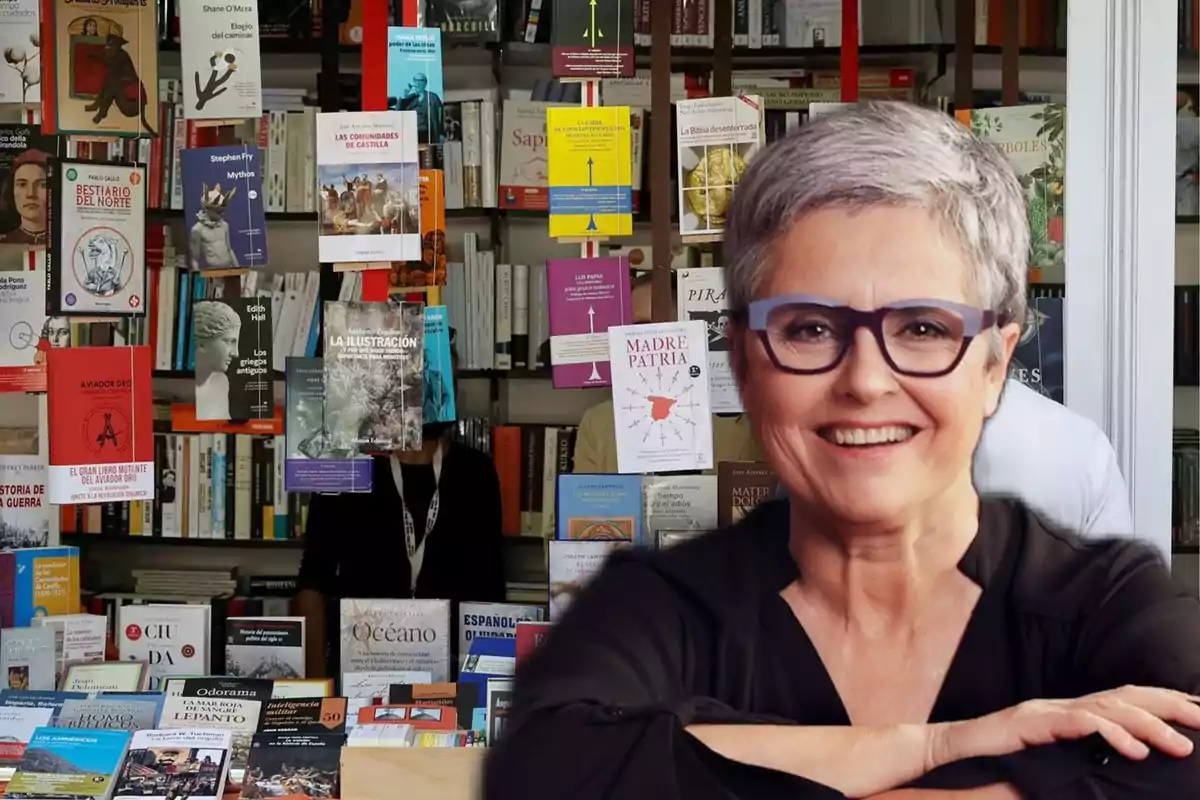 Mujer de cabello corto y canoso con gafas sonríe frente a una librería llena de libros exhibidos en estantes y colgados