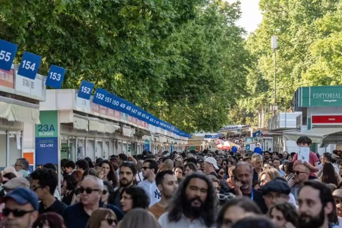 Multitud de personas caminando entre casetas de una feria de libros al aire libre rodeada de árboles Multitud de personas caminando entre casetas de una feria de libros al aire libre rodeada de árboles