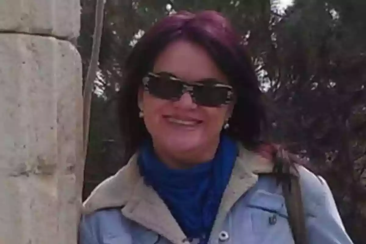 Mujer con gafas de sol y cabello oscuro sonríe al aire libre junto a una estructura de piedra Mujer con gafas de sol y cabello oscuro sonríe al aire libre junto a una estructura de piedra