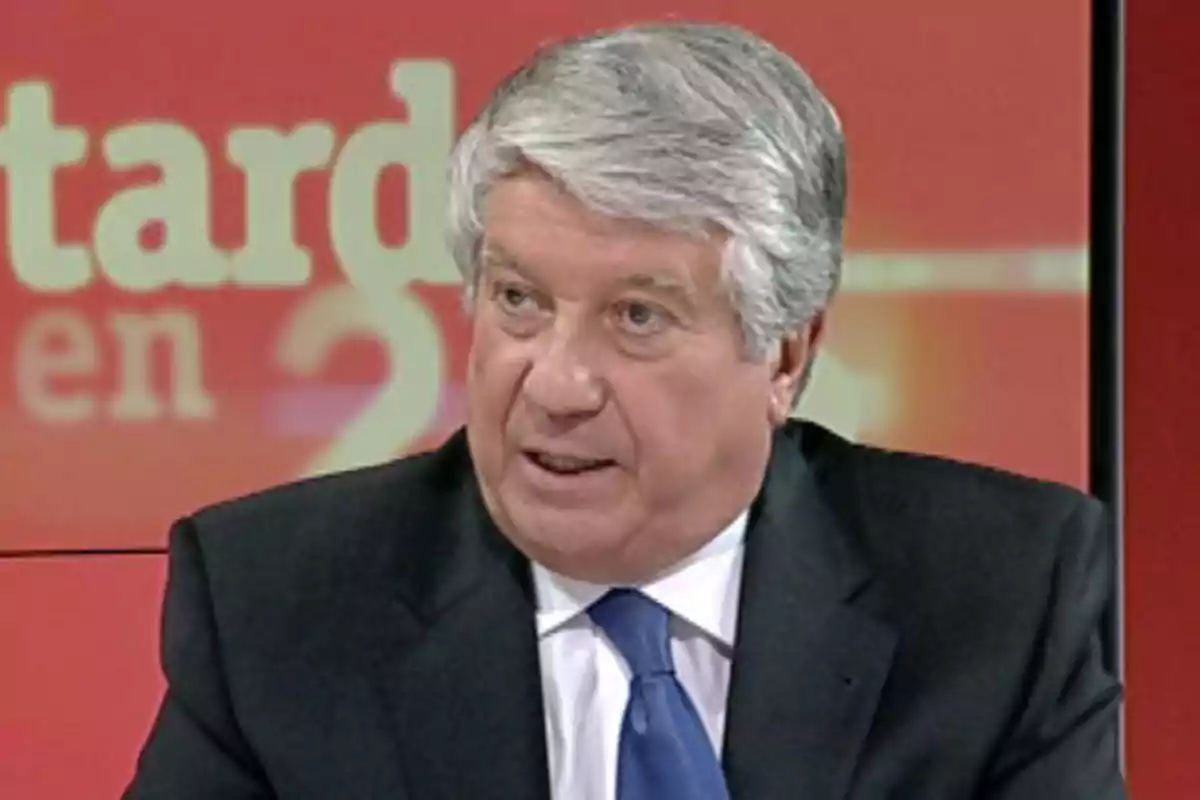Hombre de cabello canoso con traje oscuro y corbata azul hablando en un set de televisión con fondo rojo y letras desenfocadas Hombre de cabello canoso con traje oscuro y corbata azul hablando en un set de televisión con fondo rojo y letras desenfocadas