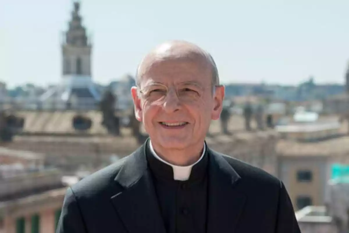 Un sacerdote sonriente con gafas y sotana negra, con un fondo urbano desenfocado.