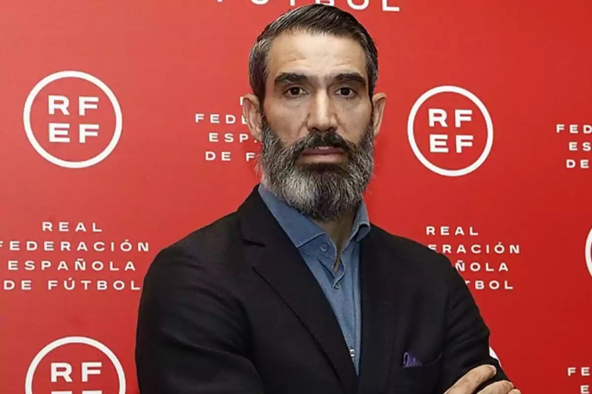 Hombre de pie con barba y traje oscuro frente a un fondo rojo con el logotipo de la Real Federación Española de Fútbol.