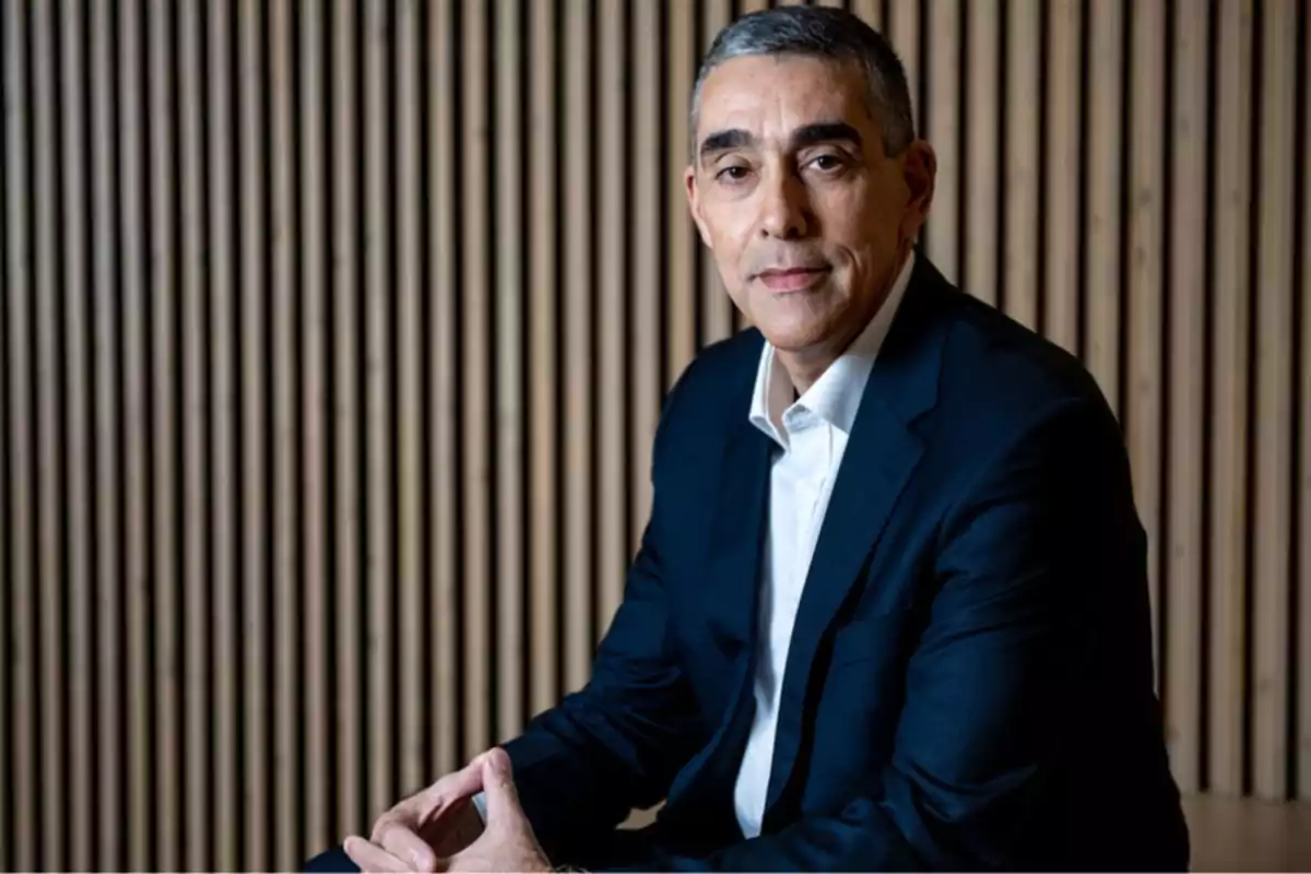Hombre de mediana edad con cabello corto y canoso vistiendo traje oscuro y camisa blanca sentado frente a un fondo de listones de madera Hombre de mediana edad con cabello corto y canoso vistiendo traje oscuro y camisa blanca sentado frente a un fondo de listones de madera