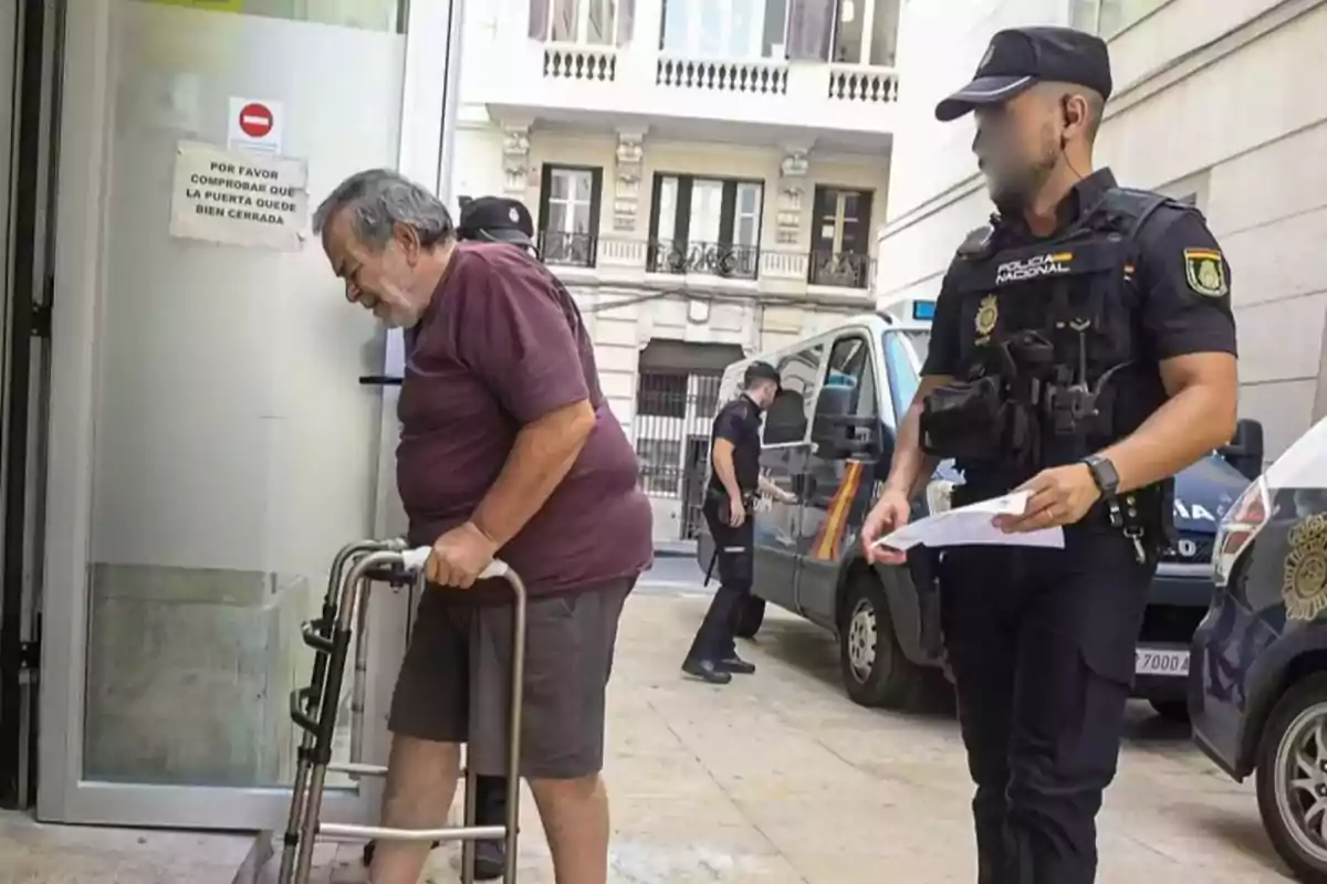 Un hombre mayor con andador entra a un edificio acompañado por un policía mientras otro agente está cerca de una furgoneta policial en la calle