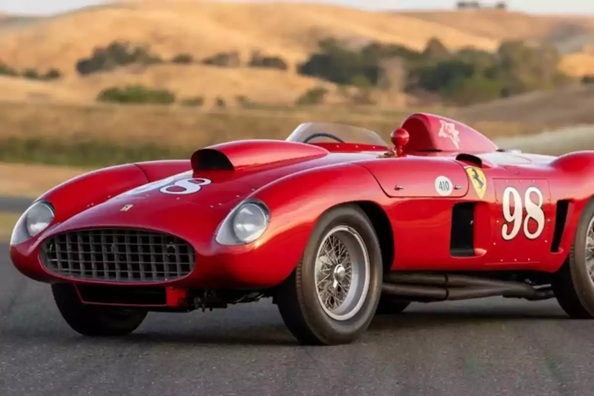 Un ferrari-410-sport-spider-by-scaglietti-1955 de carreras clásico rojo con el número 98 en un paisaje de colinas.