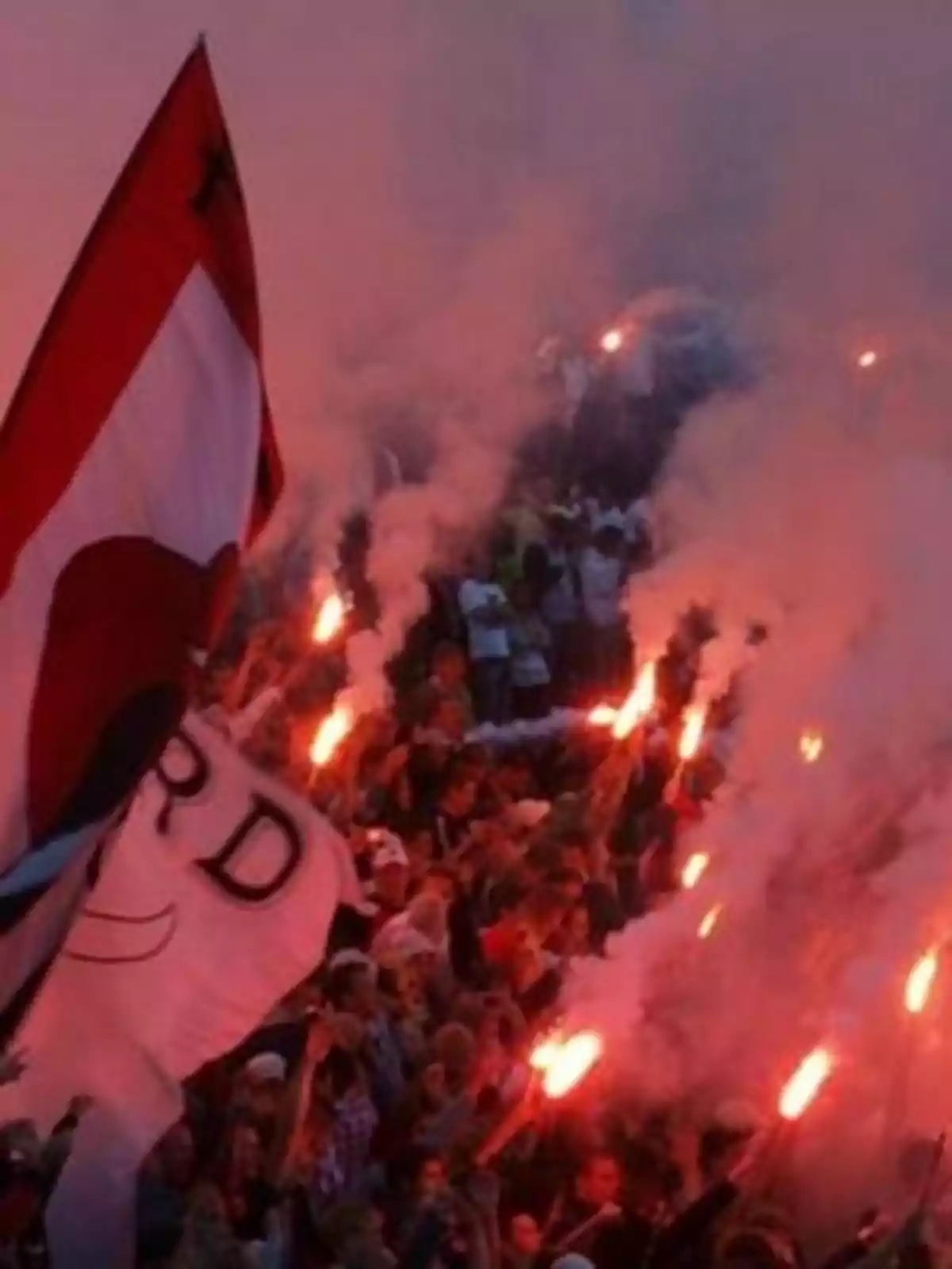 Una multitud de personas sostiene bengalas encendidas y banderas en un ambiente festivo y lleno de humo.