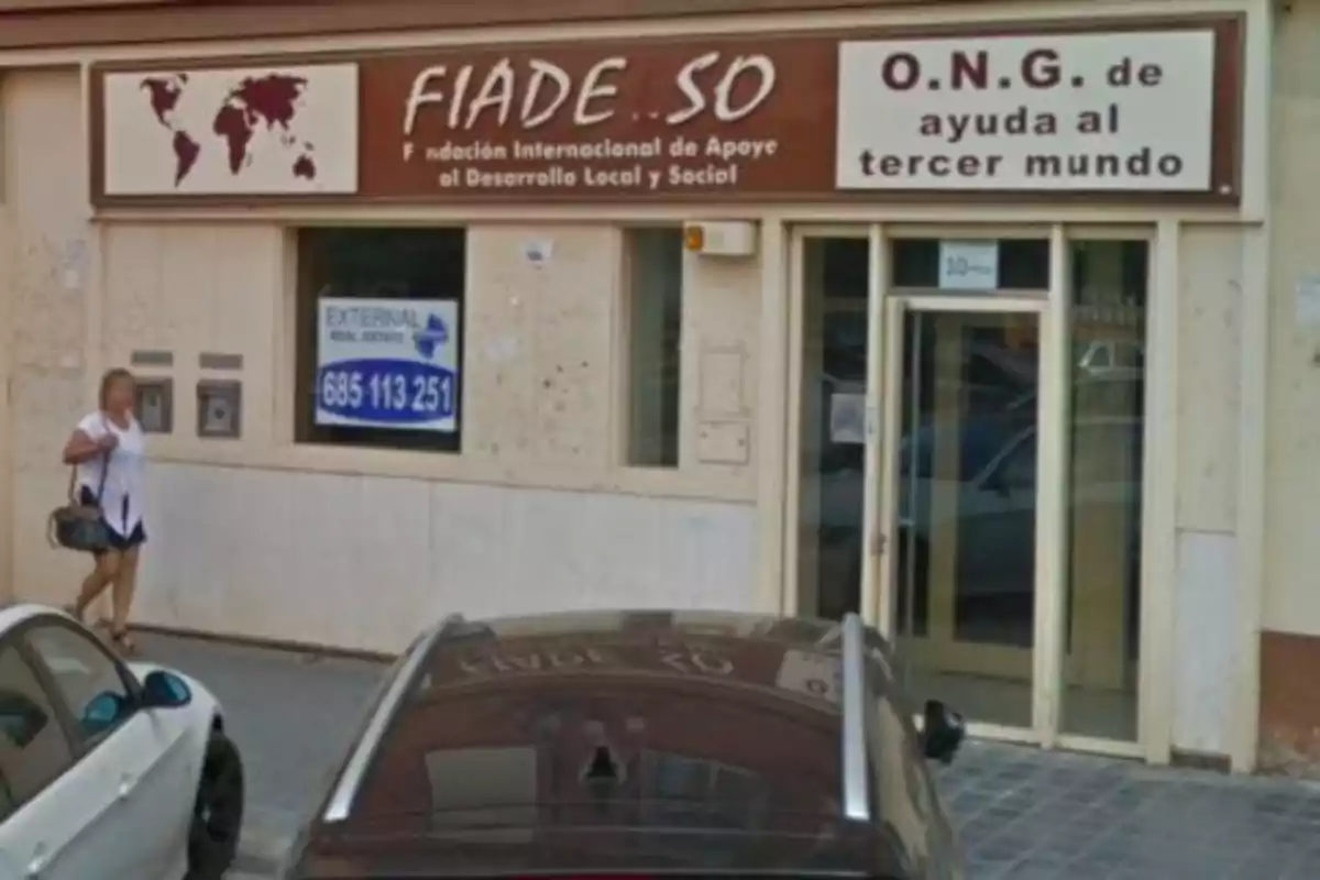 Fachada de una oficina de una ONG llamada FIADE.SO con un cartel que dice "Fundación Internacional de Apoyo al Desarrollo Local y Social" y "O.N.G. de ayuda al tercer mundo", con una persona caminando por la acera y varios coches estacionados.