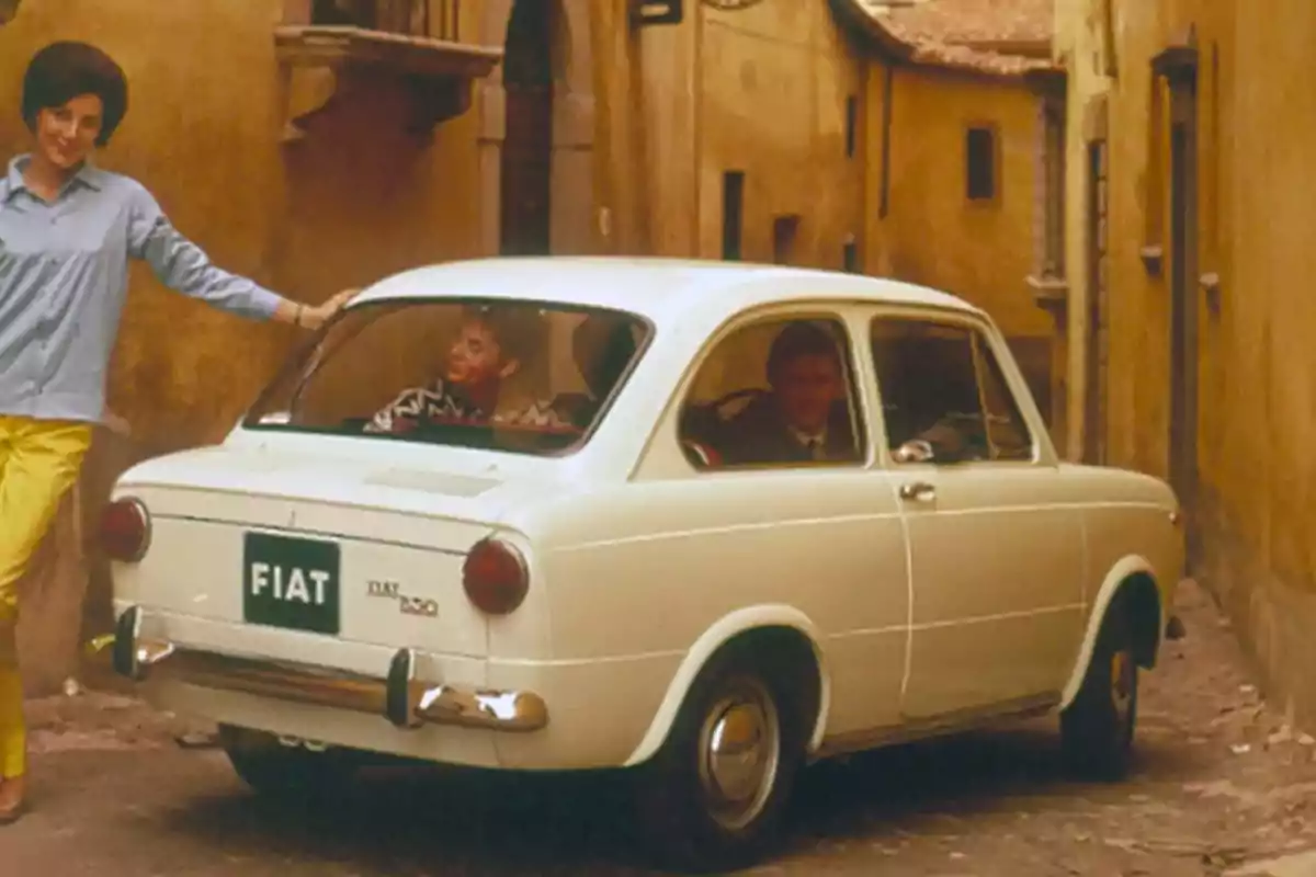 Un coche FIAT 850 blanco en una calle estrecha con una persona apoyada en el vehículo y dos personas dentro.