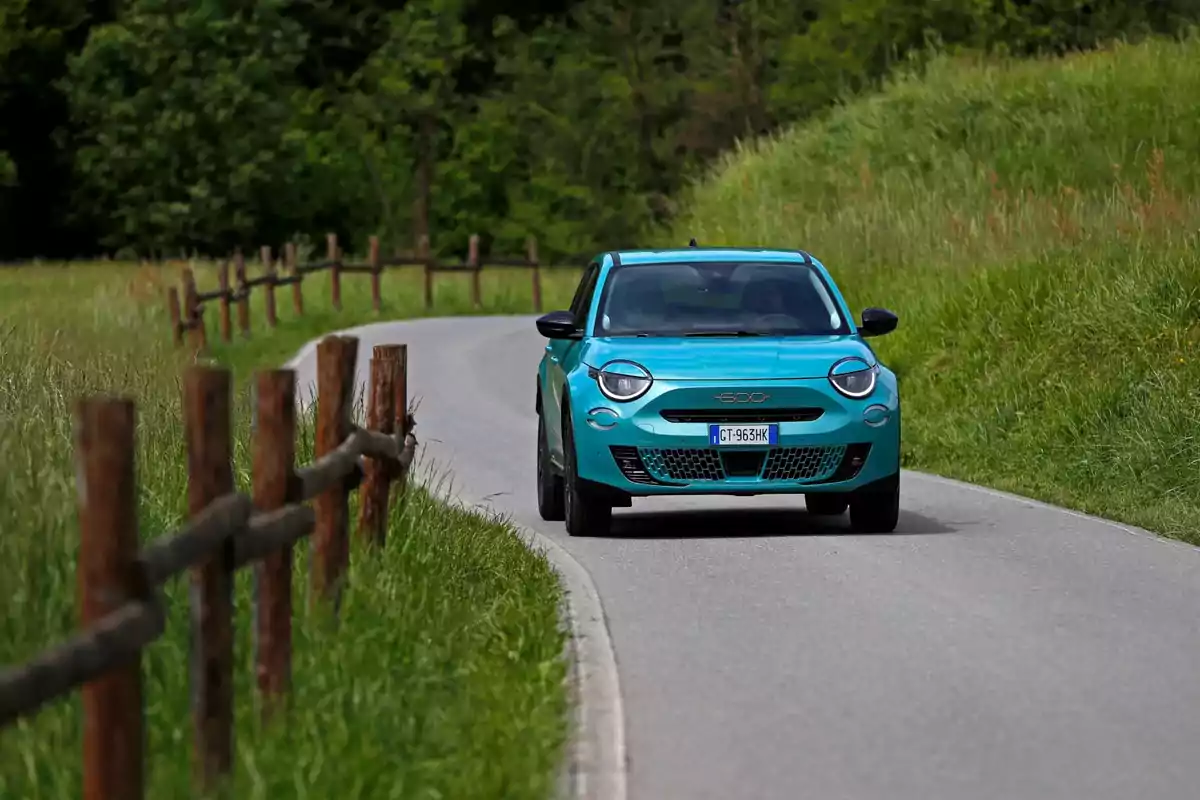 Un FIAT 600 Hybrid azul circula por un camino rural rodeado de vegetación y una cerca de madera.