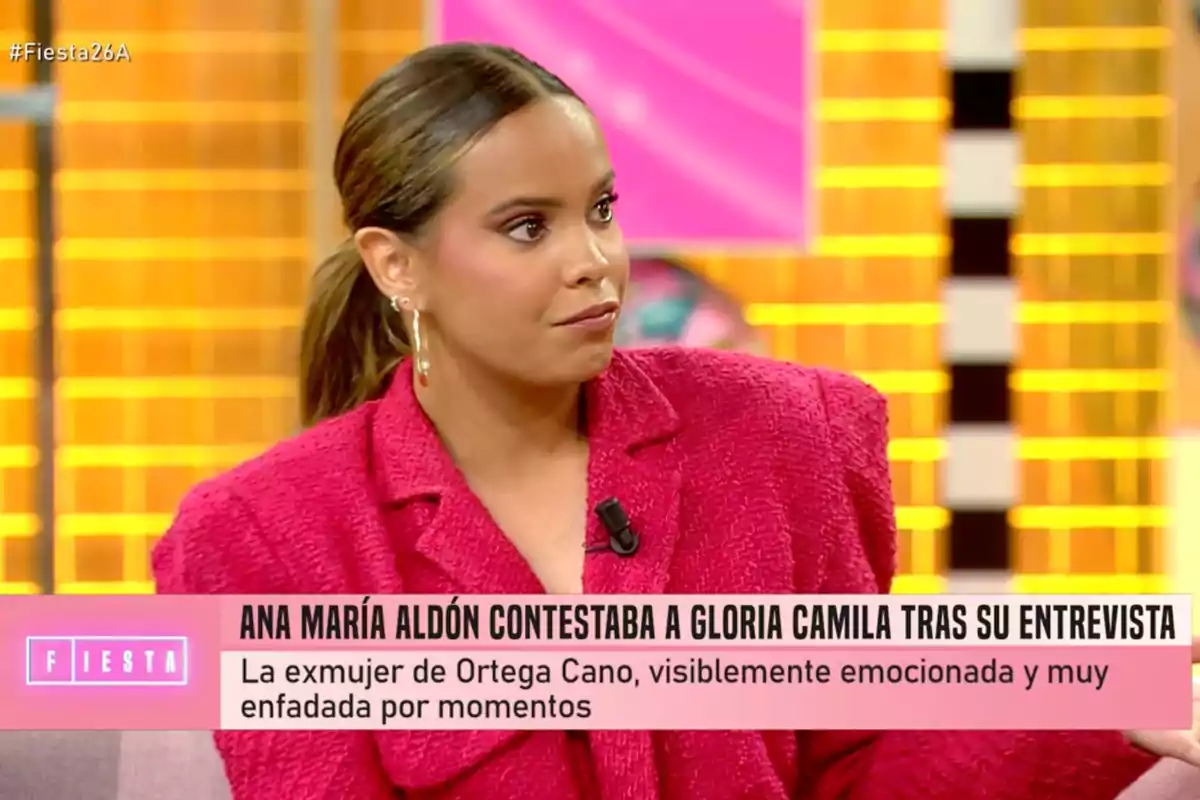 Gloria Camila con chaqueta roja y pendientes grandes está sentada en un plató de televisión con un fondo colorido, mientras un texto en la parte inferior menciona una respuesta a una entrevista. Captura de 'Fiesta' Gloria Camila con chaqueta roja y pendientes grandes está sentada en un plató de televisión con un fondo colorido, mientras un texto en la parte inferior menciona una respuesta a una entrevista. Captura de 'Fiesta'